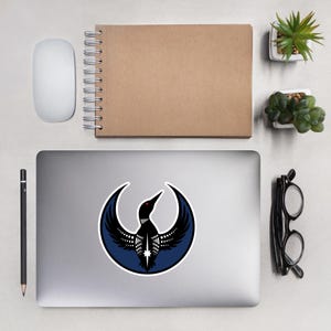May include: A laptop sticker featuring a stylized black loon with a red eye, set against a dark blue crescent moon design. The sticker has a white border and is placed on a laptop. Other items include a mouse, notebook, and pencil.