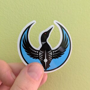 May include: A sticker depicting a black loon with spread wings, set within a light blue crescent. The loon has a red beak and white dot details. The sticker is held against a light green background.