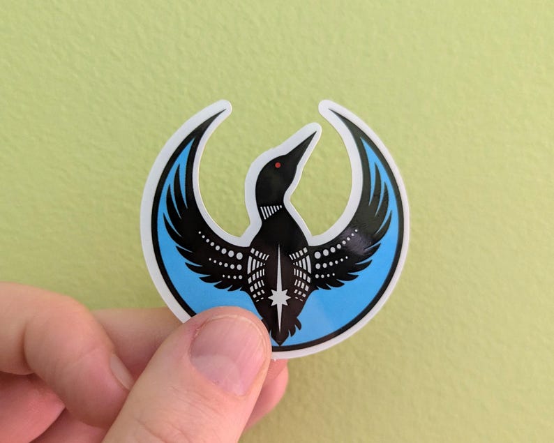 May include: A sticker depicting a black loon with spread wings, set within a light blue crescent. The loon has a red beak and white dot details. The sticker is held against a light green background.