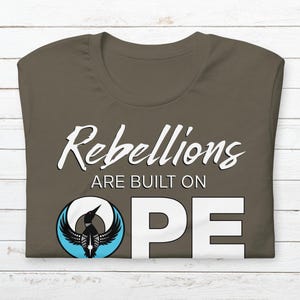 May include: A dark olive green t-shirt with the words "Rebellions are built on HOPE" in white script. The word "HOPE" is in large, bold, white capital letters. A circular graphic of a bird with outstretched wings is on the shirt.
