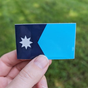 Puede incluir: Una bandera azul y negra con una estrella blanca de siete puntas en la esquina superior izquierda. La bandera está hecha de vinilo y tiene un acabado brillante.