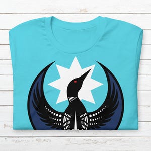 May include: A folded, light blue t-shirt with a graphic of a black bird with outstretched wings and a white starburst. The bird has a red eye and white dotted accents. The shirt is made of soft material.