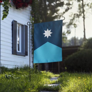 May include: A rectangular garden flag with a dark blue upper section and a light blue lower section. A white eight-pointed star is centered on the dark blue area. The flag is hanging on a black metal stand in a grassy yard.