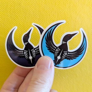 Puede incluir: Dos pegatinas con un diseño estilizado de un somorgujo. Los somorgujos son negros con detalles blancos y ojos rojos, sobre una forma de media luna. Una pegatina tiene un fondo azul oscuro, la otra un fondo azul claro. Las pegatinas están sobre un fondo amarillo.