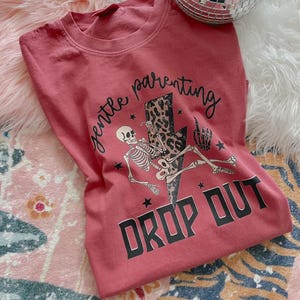 May include: Red t-shirt with a skeleton graphic holding a drink and sitting on a lightning bolt with the text "gentle parenting drop out".