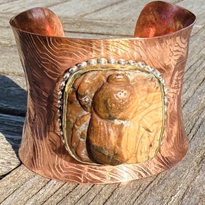 May include: A wide copper cuff bracelet with a textured surface and a central rectangular stone. The stone is a light brown color with a carved design. The bracelet has a silver-colored border around the stone.
