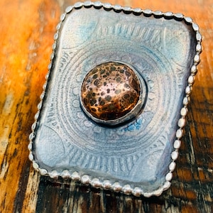 May include: A rectangular ring featuring a dark, oxidized silver-toned base with an embossed circular pattern. The center is set with a round, copper-colored cabochon displaying a speckled, dark brown texture, framed by a beaded silver edge.