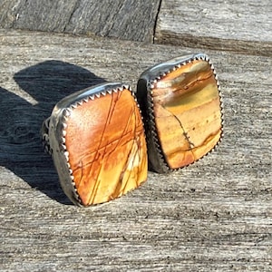 May include: Two square-shaped rings with natural stone faces. The stones have a mix of orange, brown, and yellow hues with dark vein-like patterns. The rings are set in silver-colored metal with a decorative, serrated edge.