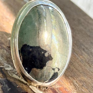 May include: A silver ring featuring a large, oval-shaped stone with a smooth, polished surface. The stone displays a mix of metallic gold, grey, and deep black patches, set within a simple, polished silver bezel that frames the oval gemstone.