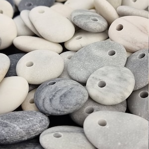 Pode incluir: Uma coleção de pedras lisas, brancas e cinzas com furos perfurados. As pedras têm forma oval e têm um aspeto natural e polido.