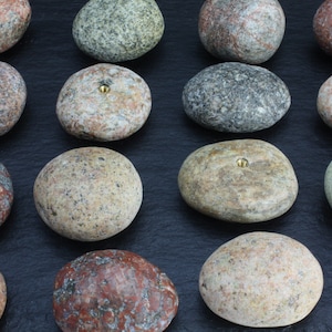 May include: A collection of 15 smooth, round, gray, brown, and green stones with a brass screw in the center of each stone.