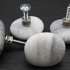 Pode incluir: Três puxadores de gaveta de pedra cinza com detalhes em metal prateado. Cada puxador tem um parafuso e um anel de metal decorativo.