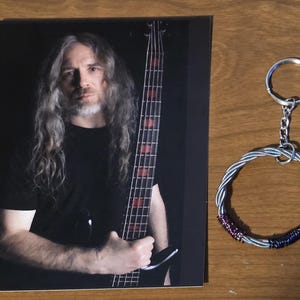 Può includere: Una fotografia di un uomo con lunghi capelli grigi che tiene un basso. Un portachiavi color argento con un design a filo ritorto e un portachiavi è accanto alla foto. Il portachiavi ha accenti di filo viola e blu.
