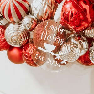 Peut inclure: Ornement de Noël rond et transparent avec un fond rouge pailleté et un texte blanc qui dit "Empty Nesters Est. 2024".