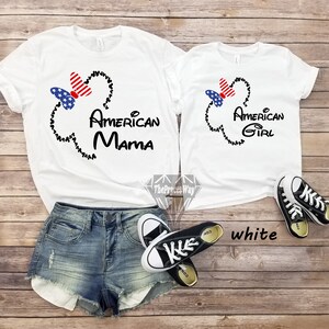 May include: Two white t-shirts with a black Mickey Mouse head outline and a red, white, and blue bow. The larger shirt says "American Mama" and the smaller shirt says "American Girl".