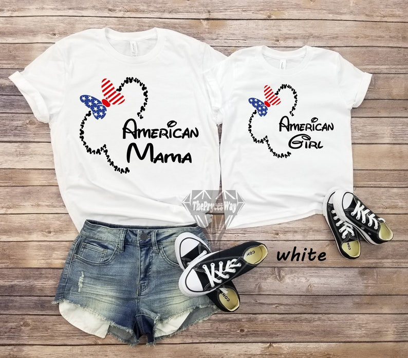 May include: Two white t-shirts with a black Mickey Mouse head outline and a red, white, and blue bow. The larger shirt says "American Mama" and the smaller shirt says "American Girl".
