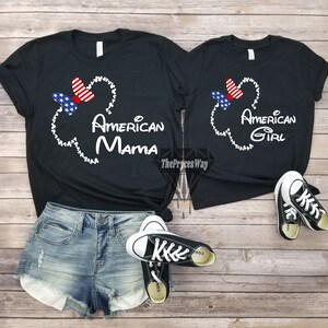 May include: Two black t-shirts with a white Mickey Mouse head outline and a red, white, and blue bow. The larger shirt says "American Mama" and the smaller shirt says "American Girl".