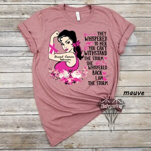 Pu&ograve; includere: Maglietta color malva con una grafica rosa di una donna con un nastro rosa, un design di fiori rosa e il testo "Breast Cancer Warrior", "They whispered to her you can't withstand the storm she whispered back I am the storm".