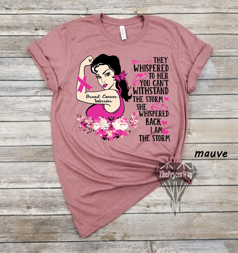 Pu&ograve; includere: Maglietta color malva con una grafica rosa di una donna con un nastro rosa, un design di fiori rosa e il testo "Breast Cancer Warrior", "They whispered to her you can't withstand the storm she whispered back I am the storm".
