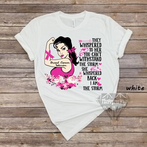 Pu&ograve; includere: Maglietta bianca con una grafica rosa di una donna con un nastro rosa, fiori e il testo "Breast Cancer Warrior" e "They whispered to her you can't withstand the storm. She whispered back I am the storm."