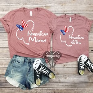 May include: Two mauve t-shirts with a white Mickey Mouse head outline and a red, white, and blue bow. The larger shirt says "American Mama" and the smaller shirt says "American Girl".