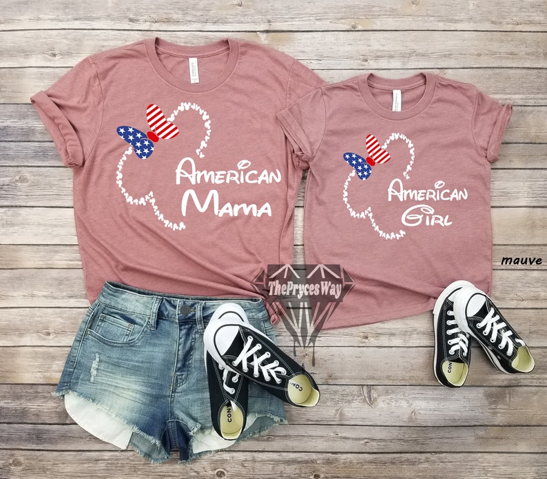 May include: Two mauve t-shirts with a white Mickey Mouse head outline and a red, white, and blue bow. The larger shirt says "American Mama" and the smaller shirt says "American Girl".