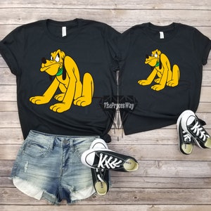 May include: Two black t-shirts with a cartoon image of Pluto the dog. The dog is yellow with a green collar. The t-shirts are on a wooden background. The text "The Prices Way" is visible on the larger t-shirt.