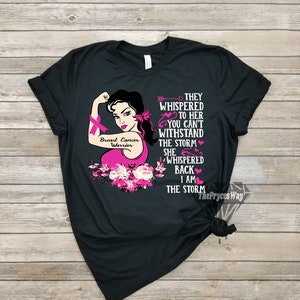 Pu&ograve; includere: Maglietta nera con un nastro rosa per la consapevolezza del cancro al seno e un'immagine di una donna con un nastro rosa sul braccio. Il testo sulla maglietta recita "Breast Cancer Warrior" e "They whispered to her you can't withstand the storm she whispered back I am the storm".