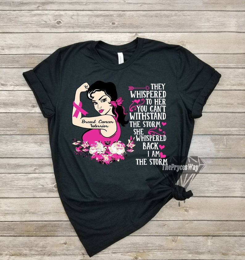 Pu&ograve; includere: Maglietta nera con un nastro rosa per la consapevolezza del cancro al seno e un'immagine di una donna con un nastro rosa sul braccio. Il testo sulla maglietta recita "Breast Cancer Warrior" e "They whispered to her you can't withstand the storm she whispered back I am the storm".