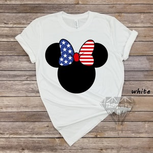 May include: White t-shirt featuring a black and white image of Mickey Mouse with a red, white and blue bow. The bow is designed with the American flag.