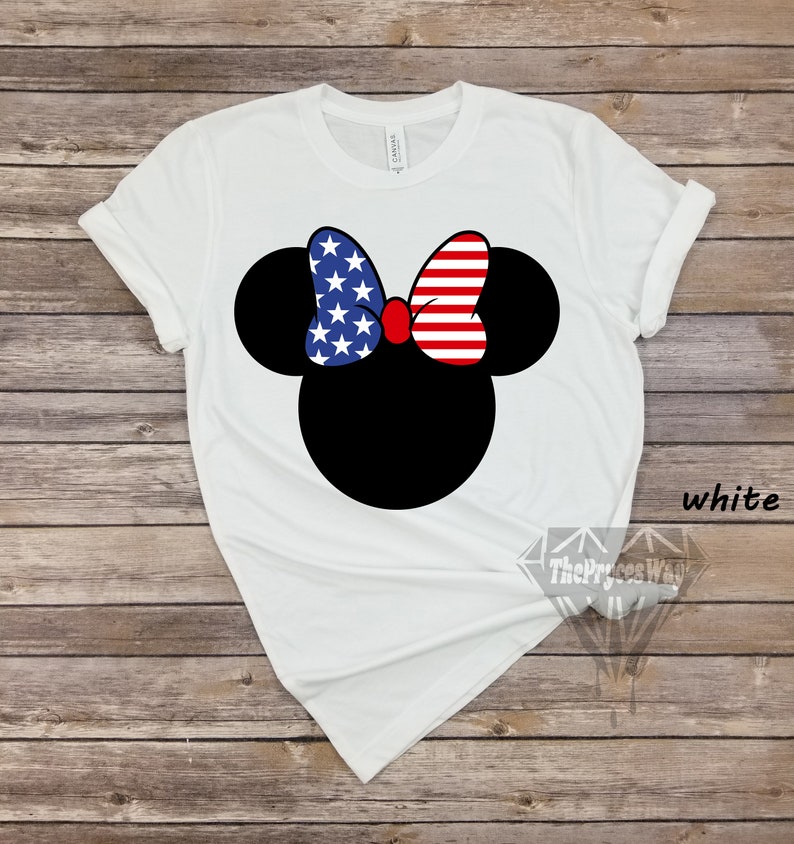 May include: White t-shirt featuring a black and white image of Mickey Mouse with a red, white and blue bow. The bow is designed with the American flag.