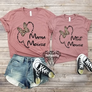 May include: Two mauve t-shirts with a black outline of Mickey Mouse's head and ears. The larger shirt says "Mama Mouse" and the smaller shirt says "Mini Mouse". Both shirts feature a leopard print bow.