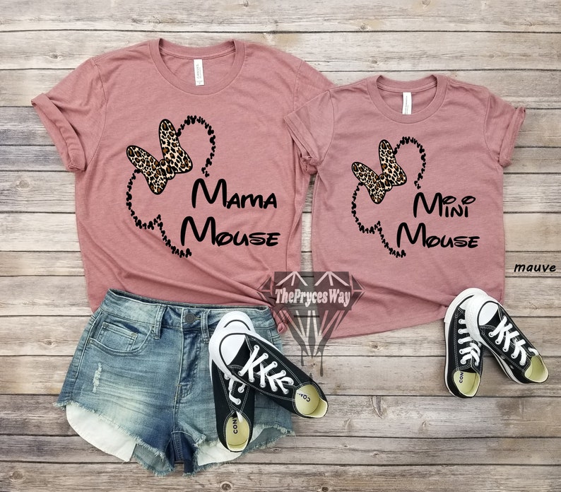 May include: Two mauve t-shirts with a black outline of Mickey Mouse's head and ears. The larger shirt says "Mama Mouse" and the smaller shirt says "Mini Mouse". Both shirts feature a leopard print bow.