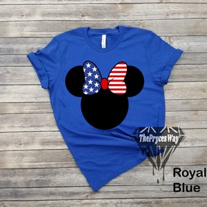 May include: A royal blue t-shirt featuring a black silhouette of Mickey Mouse wearing a red bow with an American flag pattern. The bow has white stars and red and white stripes.