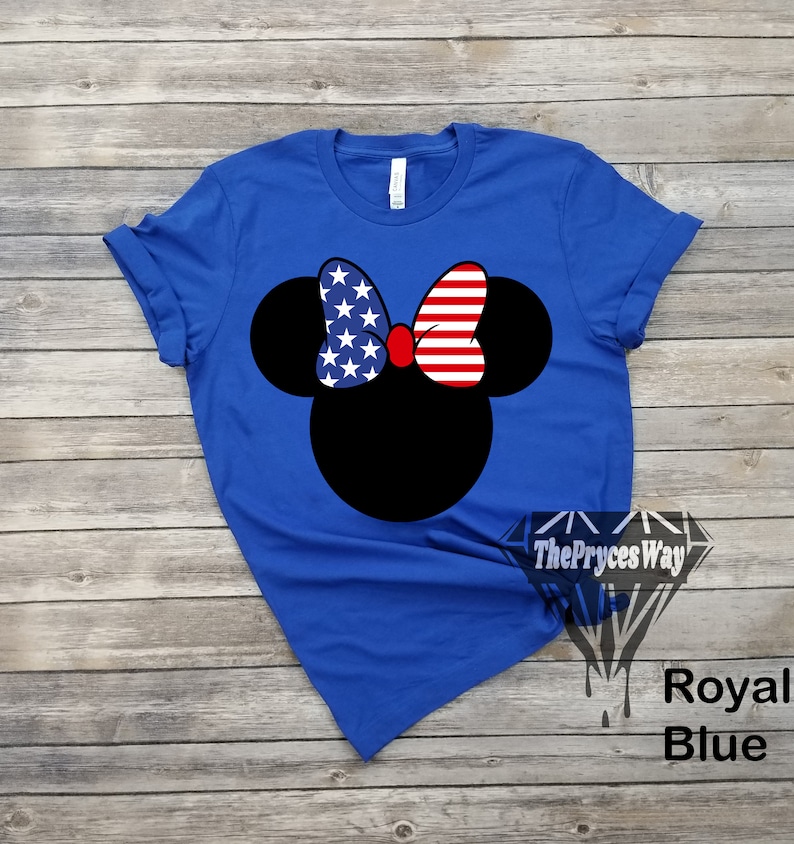 May include: A royal blue t-shirt featuring a black silhouette of Mickey Mouse wearing a red bow with an American flag pattern. The bow has white stars and red and white stripes.