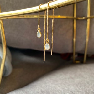 Op de afbeelding: Een paar gouden oorhangers met een traanvormige witte steen aan elke oorhanger.