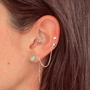 Puede incluir: Una oreja con varios piercings. La oreja está perforada con un aro con pequeñas piedras, un pequeño pendiente y un pendiente de cadena con una piedra turquesa.