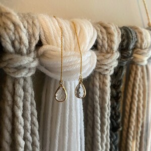 Op de afbeelding: Een paar delicate gouden oorbellen met heldere, druppelvormige kristallen. De oorbellen hangen aan fijne gouden kettingen. De achtergrond is een geweven wandkleed met verschillende tinten wit, grijs en beige garen.