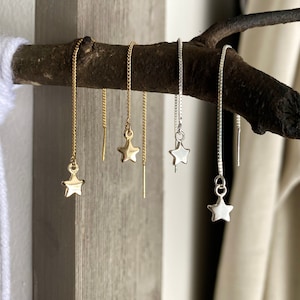 Puede incluir: Cuatro pares de pendientes colgantes con forma de estrella. Dos pares son dorados y dos plateados. Los pendientes cuelgan de delicadas cadenas y se exhiben en una rama marrón oscuro sobre un fondo neutro.