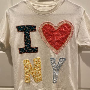 May include: A white short-sleeve t-shirt featuring a decorative applique design on the front. The design displays the text I heart NY, where the letter I is dark blue with floral patterns, the heart is red with floral details, and NY is yellow and light blue.
