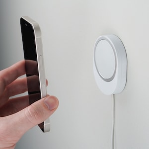 Puede incluir: Un cargador de teléfono de pared blanco y circular con un borde plateado. Un teléfono inteligente plateado se sostiene cerca del cargador. El cargador está conectado a un cable blanco. El fondo es una pared blanca lisa.
