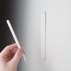 May include: A white Apple Pencil with a white magnetic wall mount. The pencil is held in a hand.
