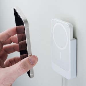 May include: A white wireless charging pad mounted on a wall with a black smartphone being held in front of it.