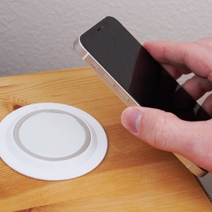 May include: A white wireless charging pad with a silver ring around the edge. A black smartphone is being held above the charging pad.