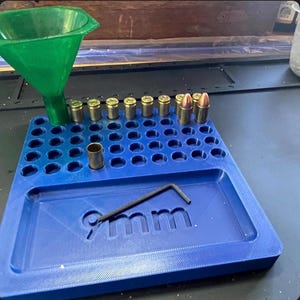 Puede incluir: Una bandeja de carga de plástico azul con una rejilla de orificios circulares y el texto 9mm en relieve. Un embudo de plástico verde está fijado en la parte superior izquierda. Varias vainas de latón y dos cartuchos están en los orificios.