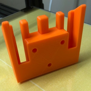 May include: Orange 3D printed plastic part with three vertical slots and three holes. The part has a rectangular shape with a raised edge on one side.