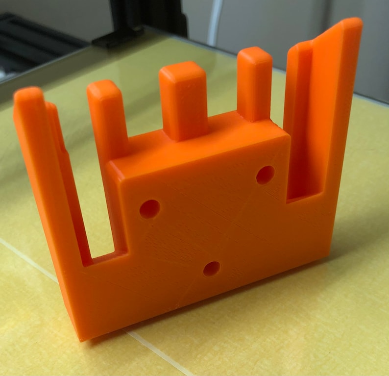 May include: Orange 3D printed plastic part with three vertical slots and three holes. The part has a rectangular shape with a raised edge on one side.