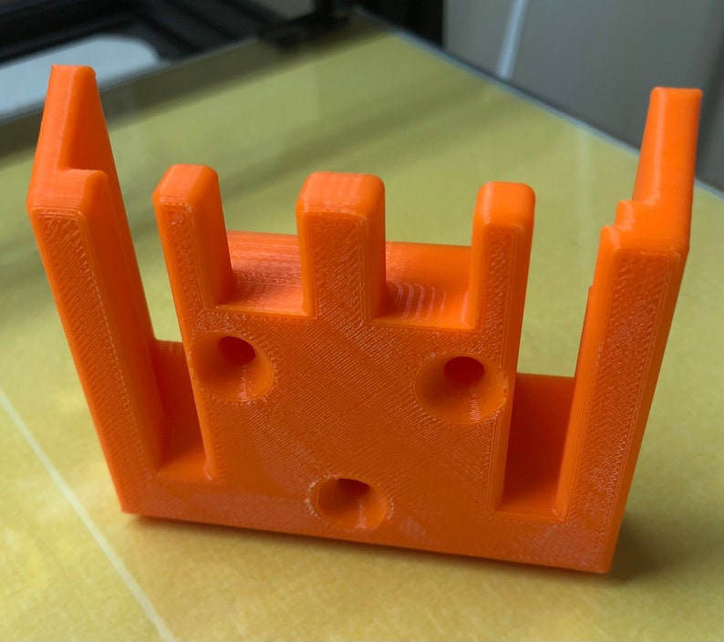May include: Orange plastic 3D printed object with four vertical slots and three circular holes.