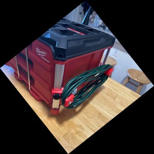 May include: A red and black Milwaukee Packout tool storage unit featuring a black top lid and silver metal corner supports. A coiled green extension cord is secured to the side of the unit using two red plastic mounting brackets attached to the frame.