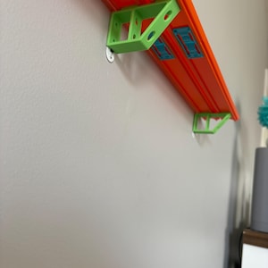 Puede incluir: Una pista naranja de Hot Wheels montada en una pared gris con soportes verdes y azules. La pista se sujeta con soportes verdes y conectores azules. La pista está diseñada para coches de juguete.